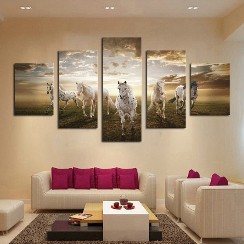 Beautiful Running Horses wall art showing five majestic white horses galloping across a grassland at sunset, displayed as 5-piece canvas panel set above modern white sofa with pink cushions