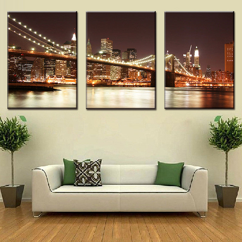 The Brooklyn Bridge at Night - Stunning 3-panel canvas wall art featuring illuminated Brooklyn Bridge spanning East River with glowing Manhattan skyline and city lights reflecting on water at nighttime