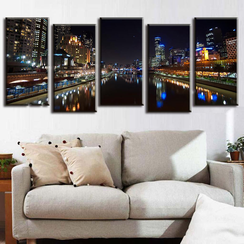 Cityscape at Night with Reflection - 5-panel canvas art showing illuminated urban buildings and skyscrapers mirrored in calm river waters, featuring glowing city lights and their reflections creating a dramatic nighttime panorama
