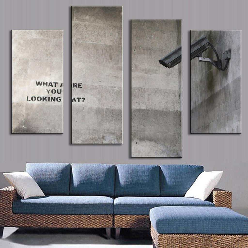 What Are You Looking At" Banksy canvas wall art featuring surveillance camera and text on concrete wall, split into 4 panels, displayed above blue sectional sofa in modern room setting