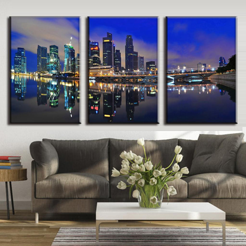 Modern City at Night - spectacular illuminated cityscape with gleaming skyscrapers reflecting in calm waterfront, 3-panel canvas wall art featuring dramatic blue twilight sky and mirror-like water reflections