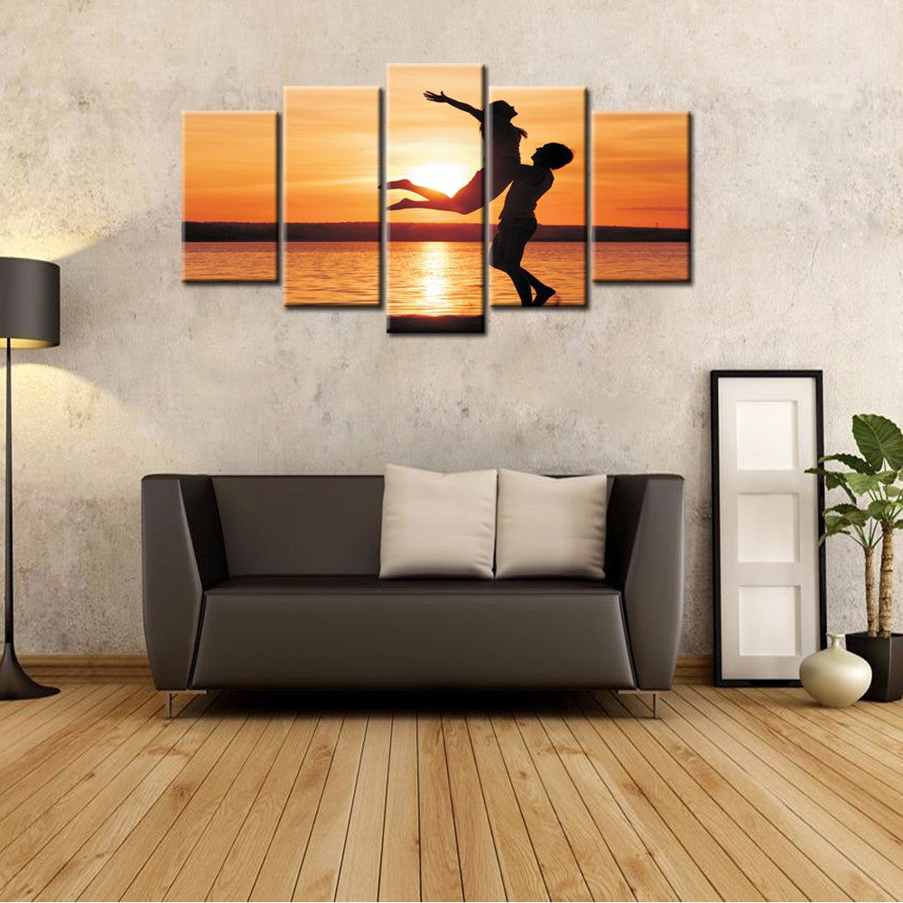Uplifted Silhouettes - 5-panel canvas wall art showing two silhouetted figures in graceful lift pose against vibrant orange sunset over ocean, perfect for home decor and living room spaces